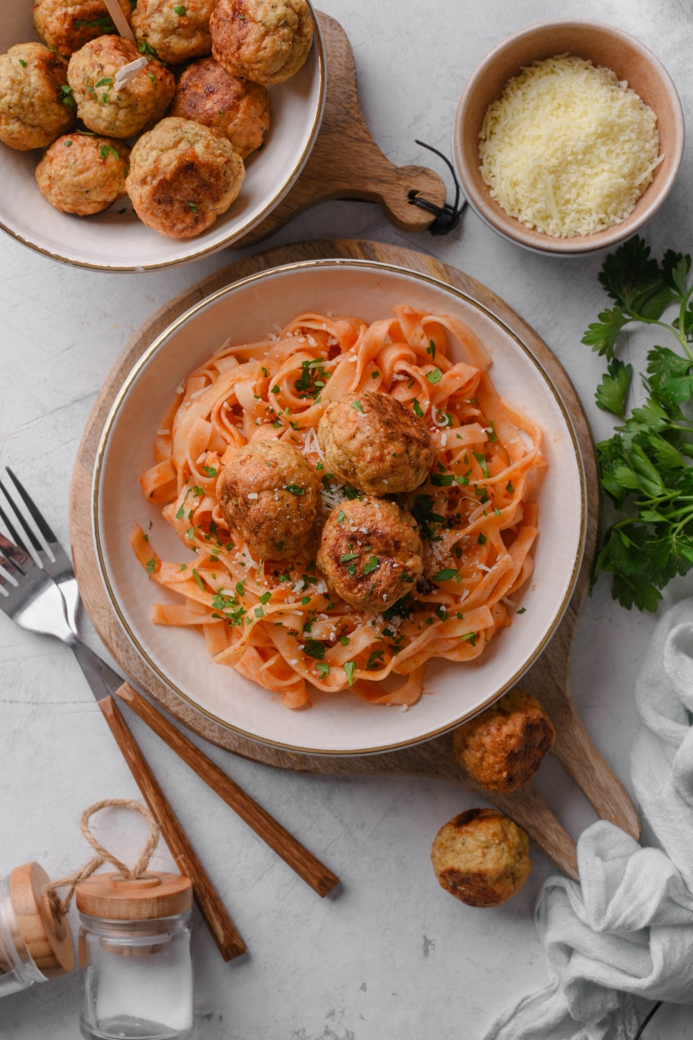 Overhead view of a plate of pasta in red sauce with three meatballs on top. The plate is surrounded by bowls of parmesan cheese and more meatballs, some of which have spilled on to the counter.