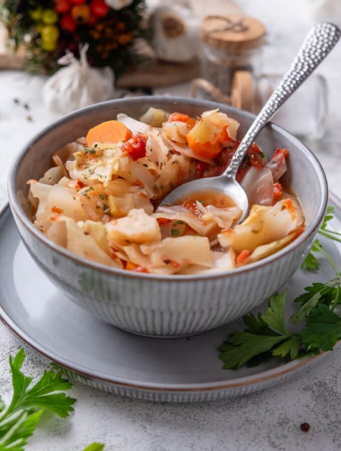 A bowl of cabbage soup with chunks of tomato, carrots, and seasonings. There is a spoon on the bowl and sprigs of cilantro next to the bowl.