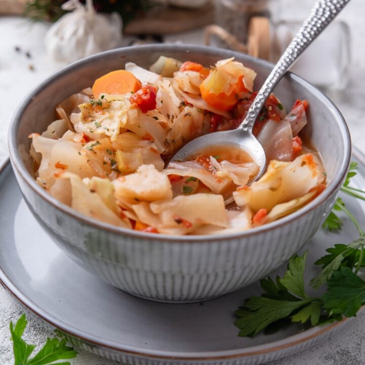 A bowl of cabbage soup with chunks of tomato, carrots, and seasonings. There is a spoon on the bowl and sprigs of cilantro next to the bowl.