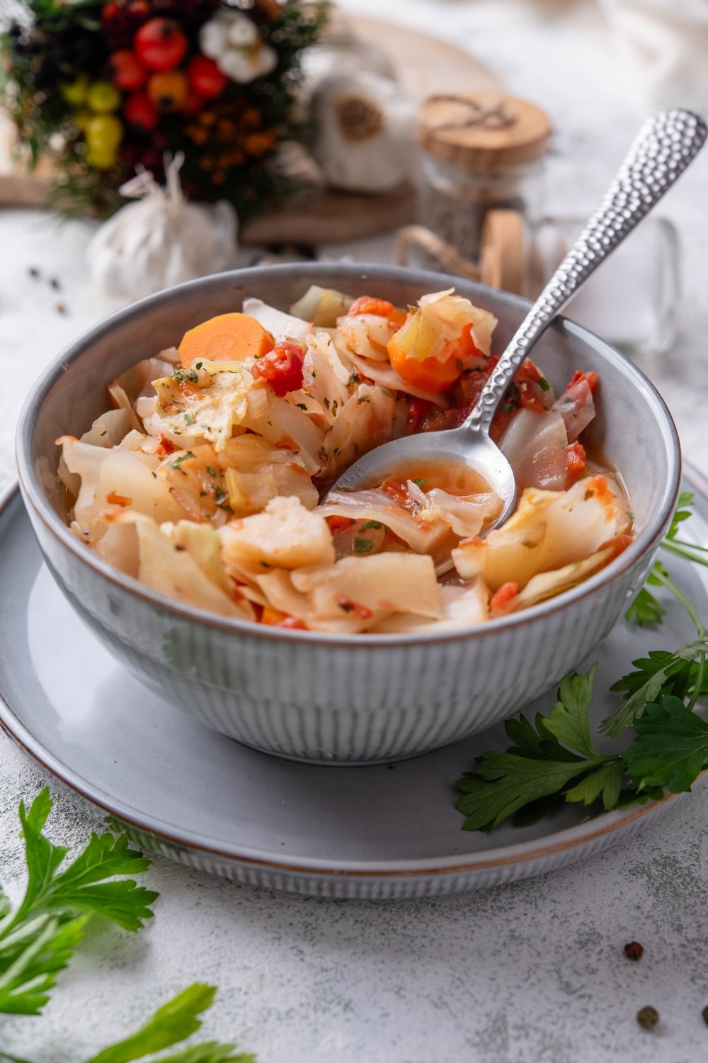 A bowl of cabbage soup with chunks of tomato, carrots, and seasonings. There is a spoon on the bowl and sprigs of cilantro next to the bowl.