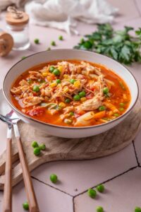 A bowl of soup with tomatoes, shredded chicken, peas, corns, and carrots.