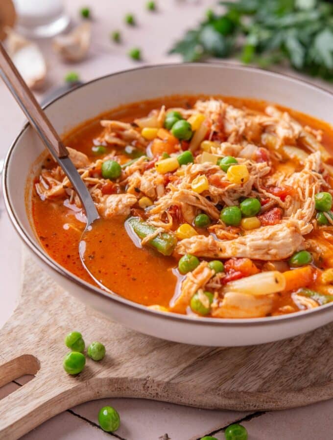 A bowl of soup with tomatoes, shredded chicken, peas, corns, and carrots. There is a spoon in the bowl.