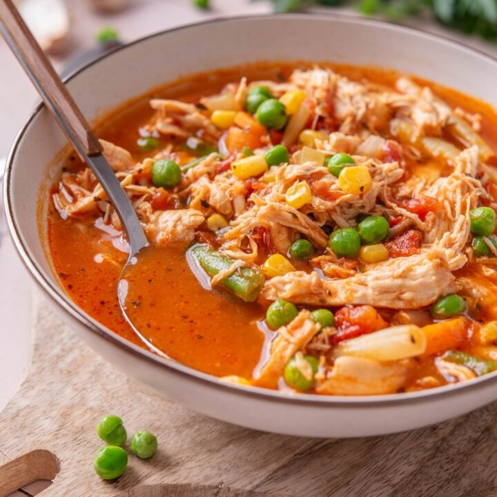 A bowl of soup with tomatoes, shredded chicken, peas, corns, and carrots. There is a spoon in the bowl.