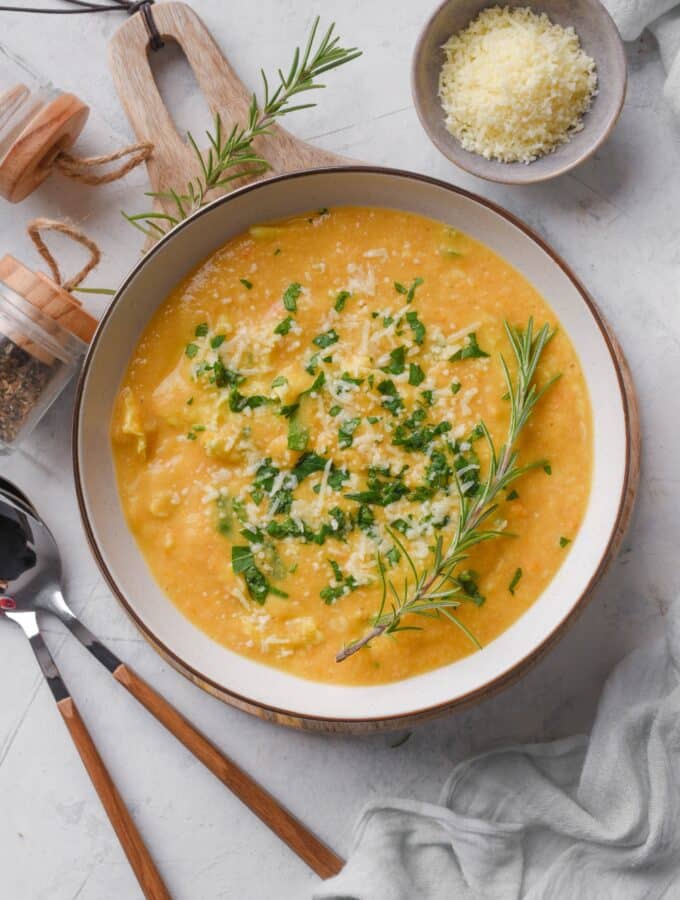 Overhead view of a bowl of creamy soup with parmesan cheese, rosemary, and kale on top. Two spoons and a bowl of parmesan cheese are next to the soup.