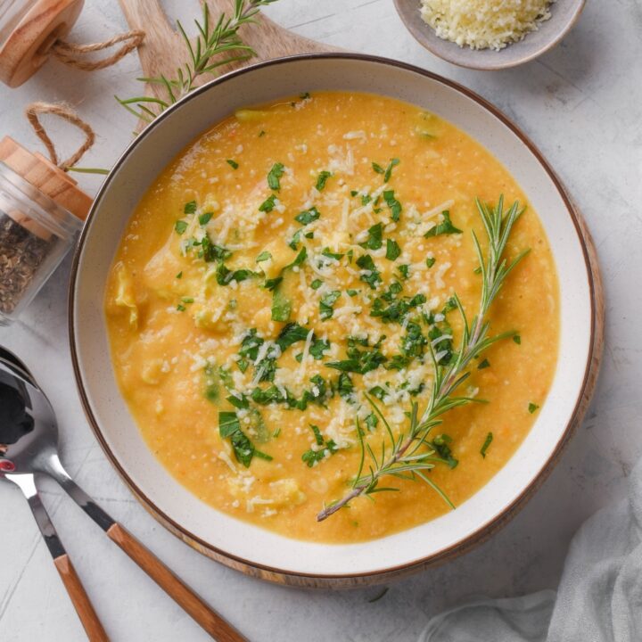 Overhead view of a bowl of creamy soup with parmesan cheese, rosemary, and kale on top. Two spoons and a bowl of parmesan cheese are next to the soup.