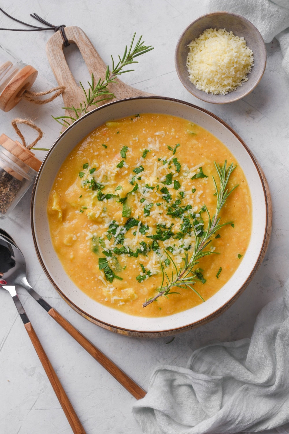 Overhead view of a bowl of creamy soup with parmesan cheese, rosemary, and kale on top. Two spoons and a bowl of parmesan cheese are next to the soup.