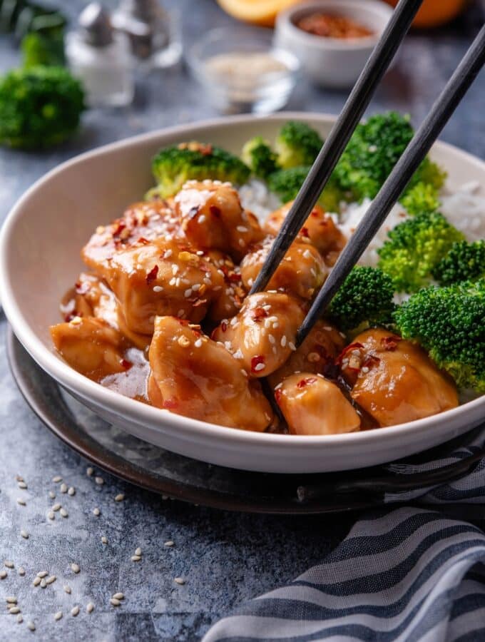 A plate of orange chicken garnished with sesame seeds and a pair of chopsticks is grabbing a piece of chicken.