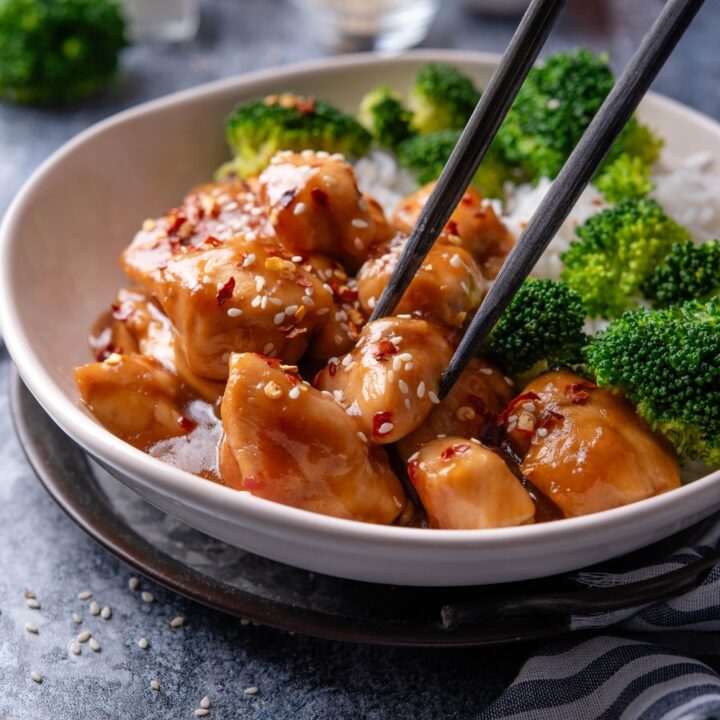 A plate of orange chicken garnished with sesame seeds and a pair of chopsticks is grabbing a piece of chicken.