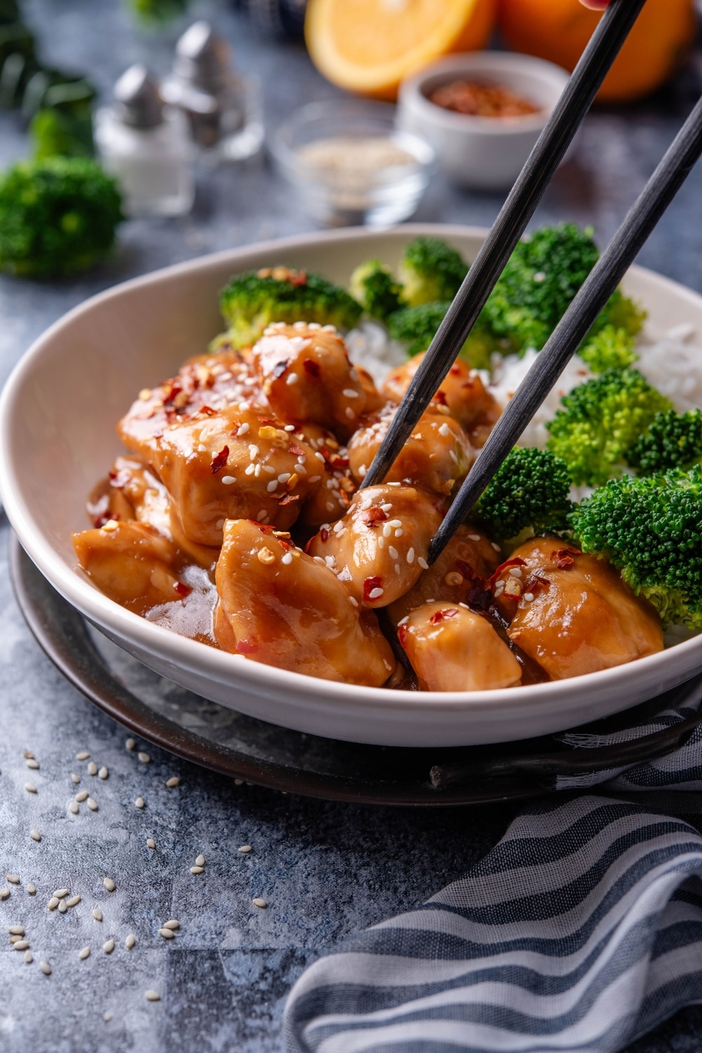 A plate of orange chicken garnished with sesame seeds and a pair of chopsticks is grabbing a piece of chicken.