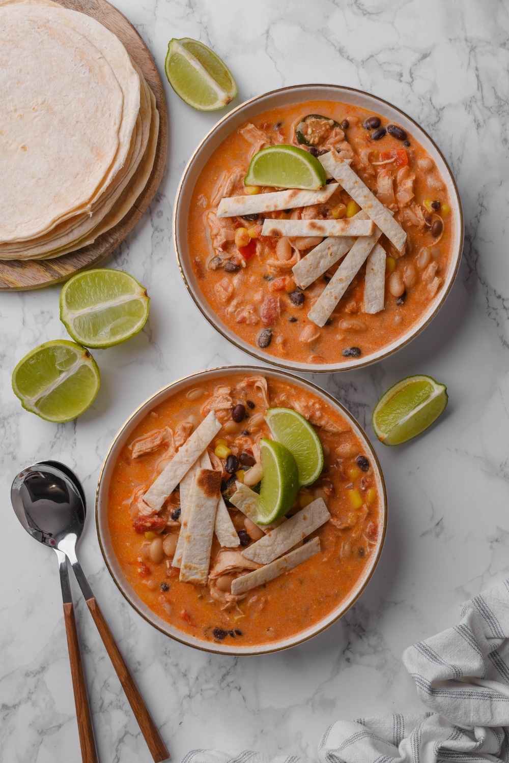 Two bowls of creamy chicken tortilla soup with corn, beans, and diced peppers, with tortilla strips and lime wedges on top.