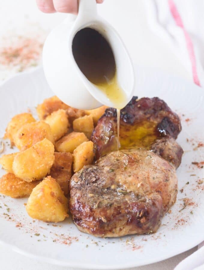 A pitcher of gravy being drizzled on a seasoned pork chop next to a side of tater tots.