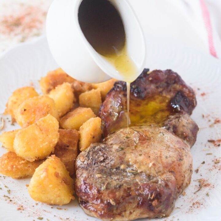 A pitcher of gravy being drizzled on a seasoned pork chop next to a side of tater tots.