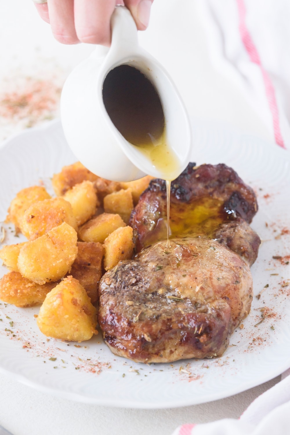 A pitcher of gravy being drizzled on a seasoned pork chop next to a side of tater tots.