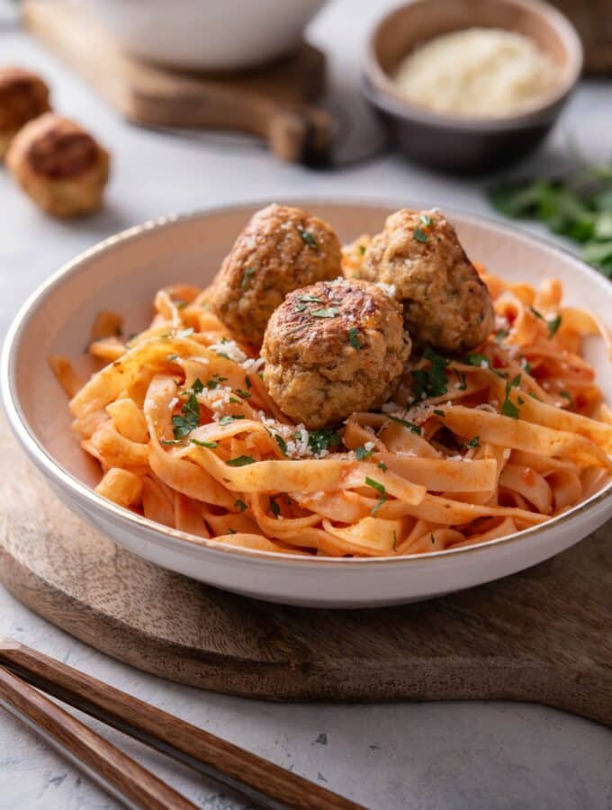 A plate of pasta in red sauce with three meatballs on top. The plate is on a wooden board next to a set of silverware.