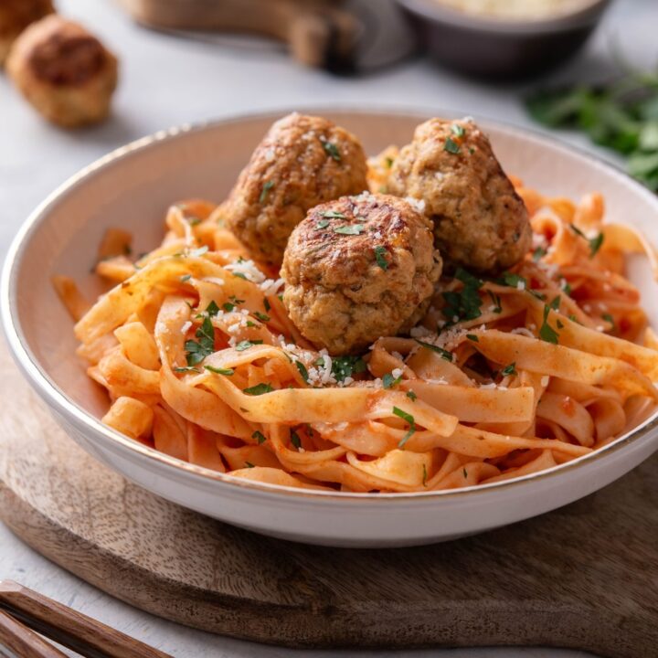 A plate of pasta in red sauce with three meatballs on top. The plate is on a wooden board next to a set of silverware.