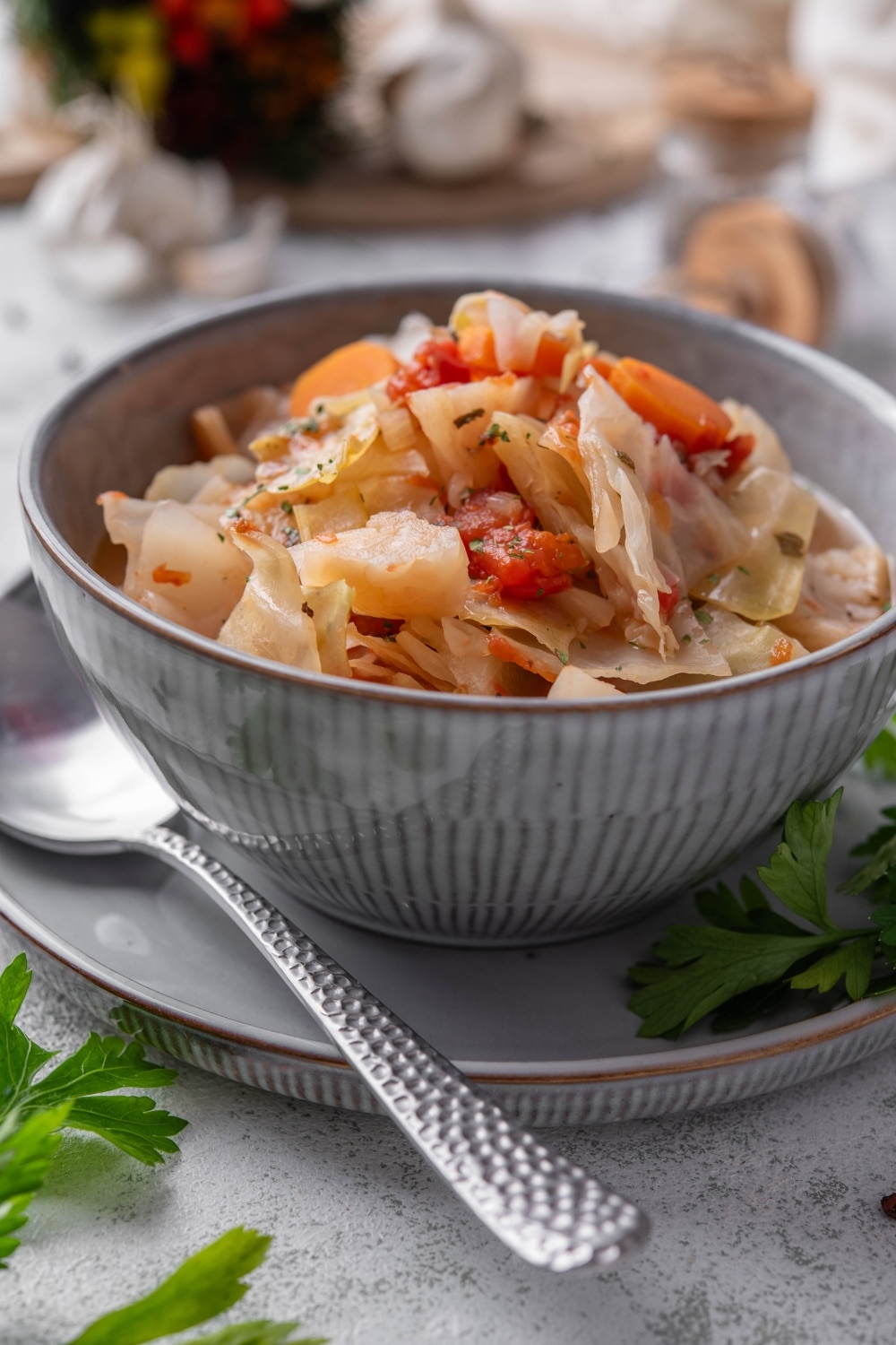 A bowl of cabbage soup with chunks of tomato, carrots, and seasonings. The bowl is on a plate next to a spoon.