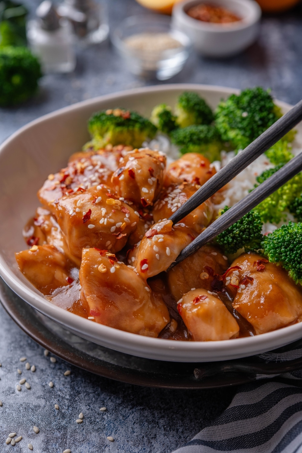 A plate of orange chicken garnished with sesame seeds and a pair of chopsticks is grabbing a piece of chicken.