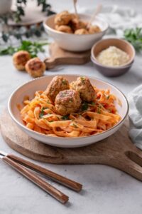 A plate of pasta in red sauce with three meatballs on top. The plate is on a wooden board next to a set of silverware.