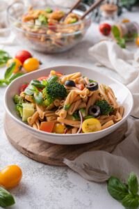 A bowl of pasta salad with black olives, cherry tomatoes, fresh herbs, cucumber, black olives, and broccoli florets.