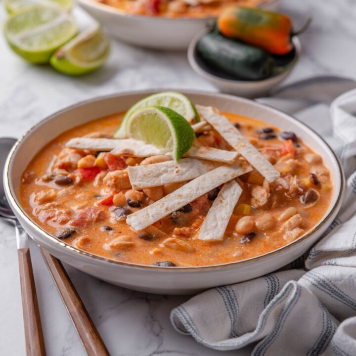 A bowl of creamy soup with diced tomatoes, corn, beans, and chicken, with crispy tortilla strips and lime wedges garnished on top.