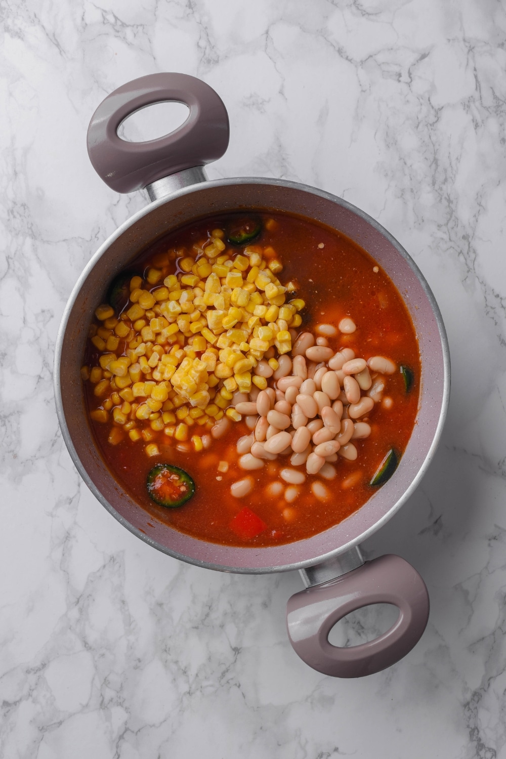 A pot filled with broth with sliced jalapeños, corn, and navy beans in it.