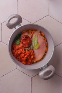 A pot filled with sliced carrots, celery, raw chicken breasts, crushed tomatoes, tomato paste, bay leaves, and seasonings.