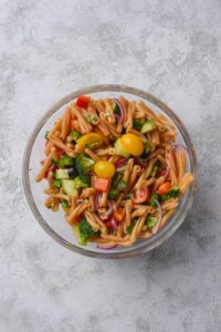 A clear bowl filled with pasta, broccoli, cherry tomatoes, red onion, peppers, and fresh herbs.
