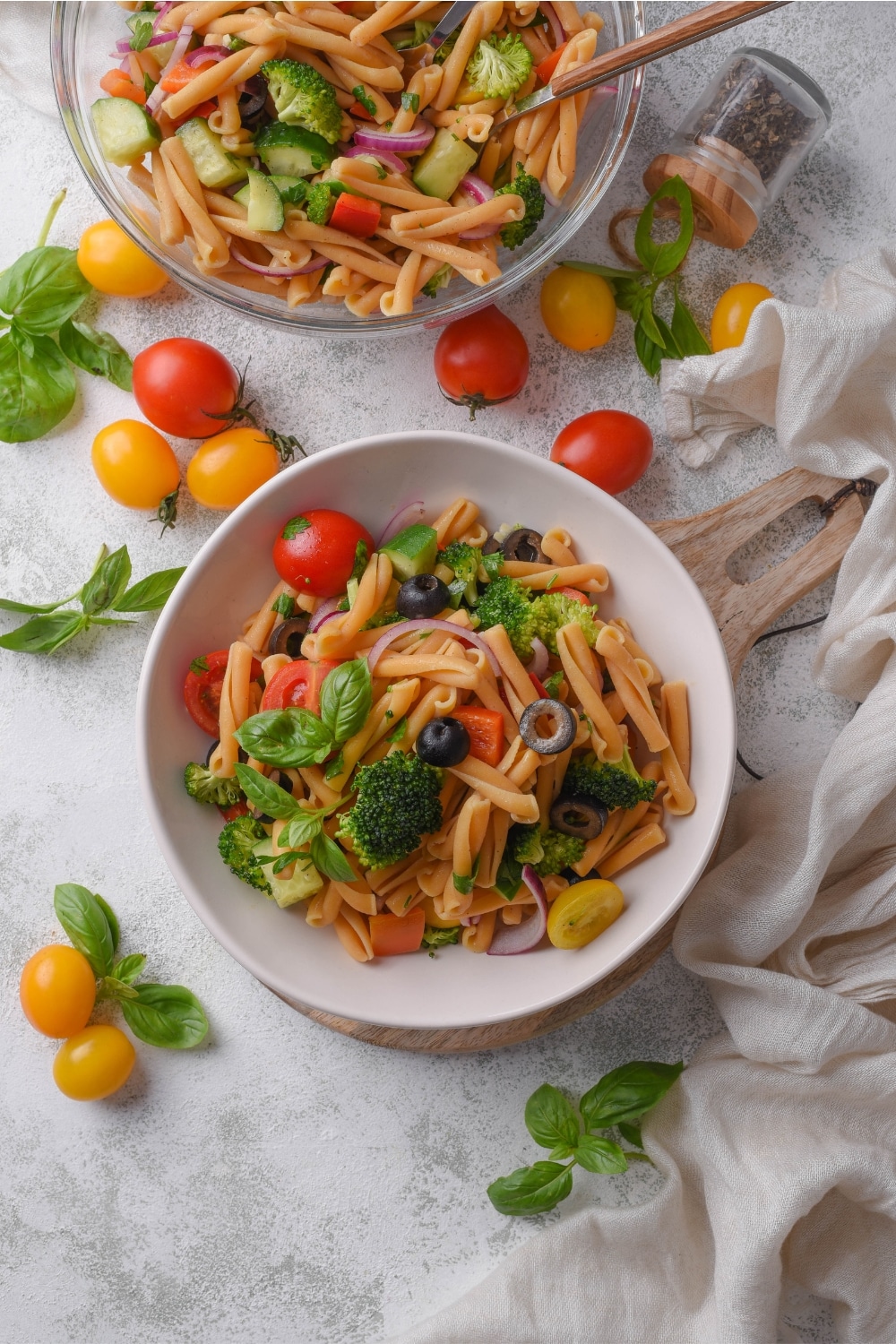 Overhead view of a bowl of pasta salad with black olives, tomatoes, basil, and broccoli. Next to the bowl is a serving bowl filled with more pasta salad and cherry tomatoes are sprinkled on the counter.