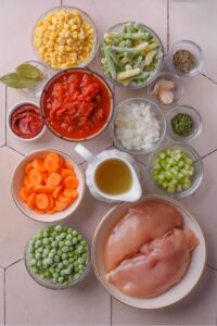 An assortment of ingredients including bowls of crushed tomatoes, corn, green and yellow beans, onion, celery, carrots, raw chicken breasts, and broth.