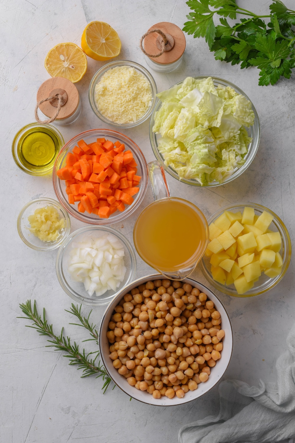 An assortment of ingredients including bowls of chickpeas, potatoes, cabbage, carrots, onion, broth, parmesan cheese, and a halved lemon, all on a white countertop.