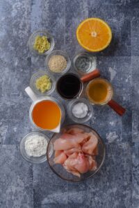 An assortment of ingredients including bowls of raw chicken, orange juice, soy sauce, vinegar, cornstarch, sesame seeds, minced ginger, and half an orange, all on a blue countertop.