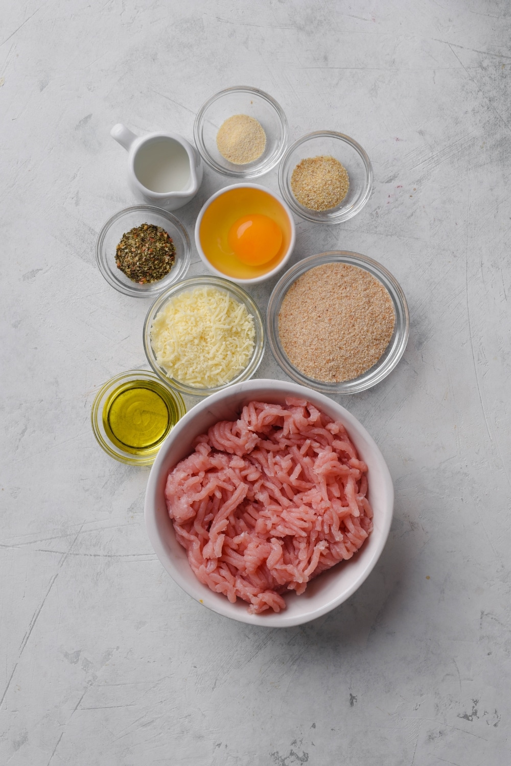 An assortment of ingredients including bowls of raw ground turkey, parmesan cheese, an egg, bread crumbs, oil, and seasonings.