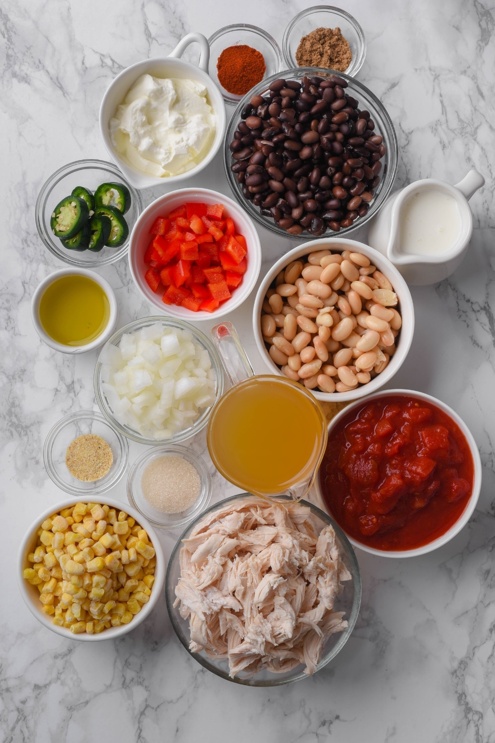 An assortment of ingredients including bowls of navy beans, black beans, crushed tomatoes, diced peppers, onion, shredded chicken, corn, heavy cream, sour cream, and spices.