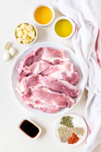 An assortment of ingredients including a plate of raw pork chops and bowls of spices, butter cubes, oil, soy sauce, and three garlic cloves.