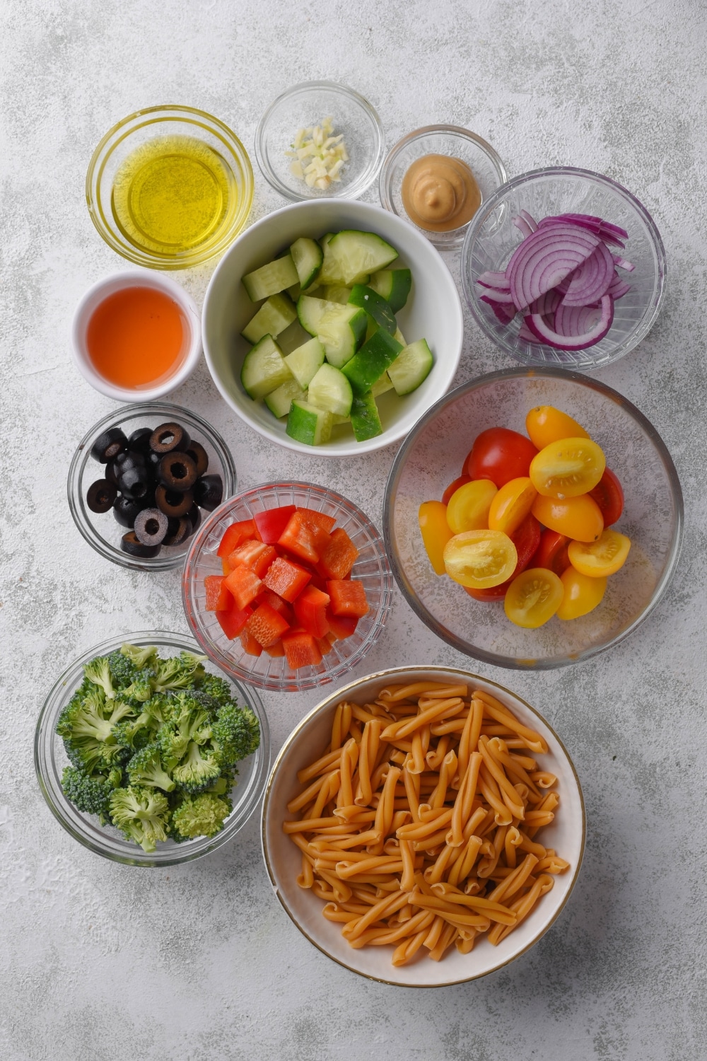 An assortment of ingredients including bowls of dried pasta, cherry tomatoes, cucumber, broccoli florets, black olives, diced peppers, sliced red onion, oil, vinegar, mustard, and minced garlic.