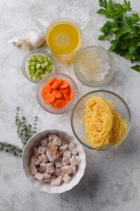 An assortment of ingredients including bowls of dried pasta, diced turkey, carrots, celery, onion, and stock.