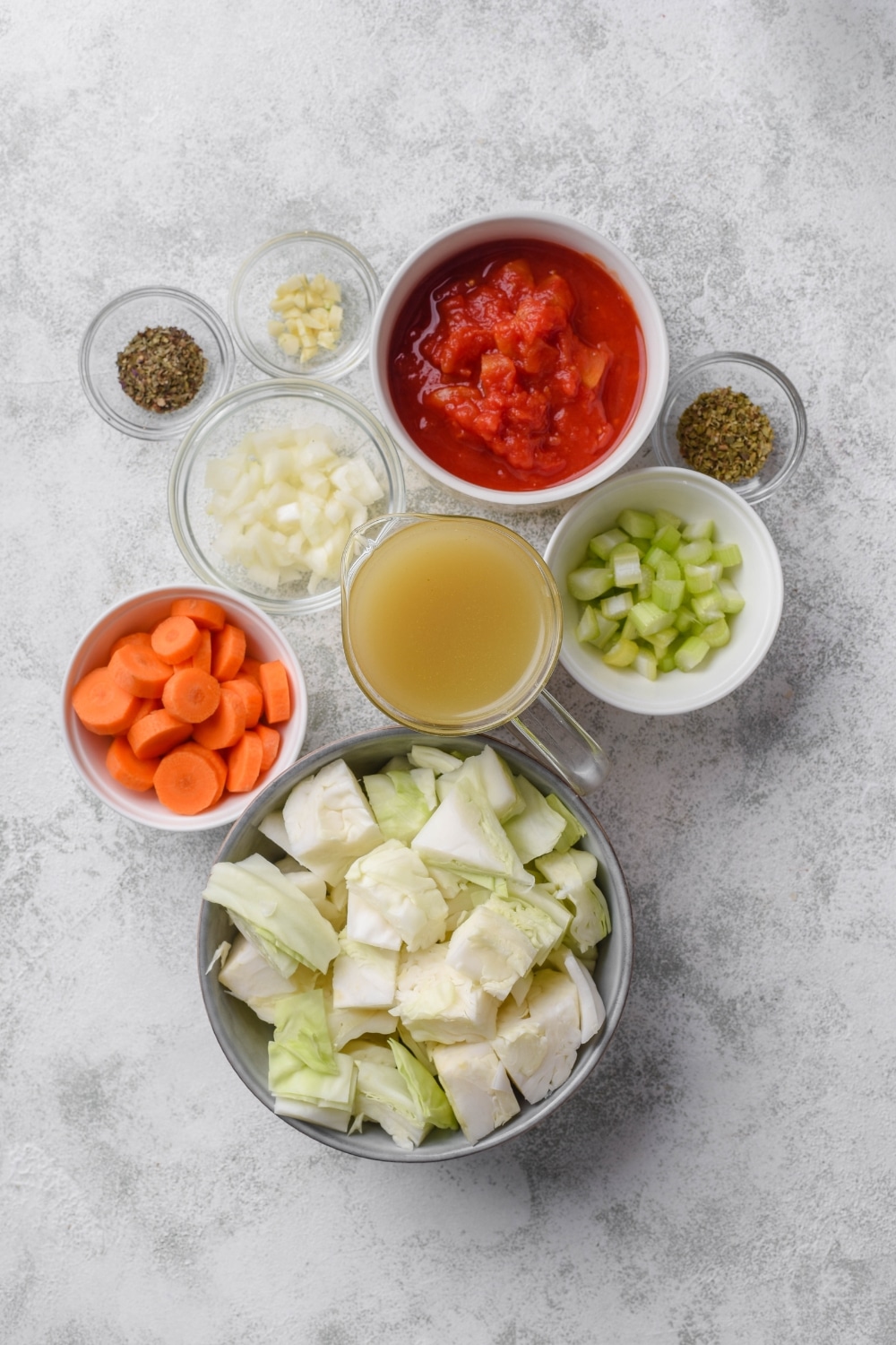 An assortment of ingredients including bowls of cabbage, celery, onion, carrots, crushed tomatoes, broth, and seasonings.