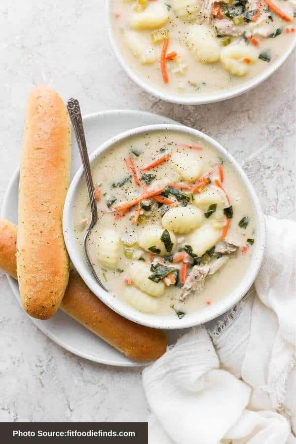 Potato gnocchi, chicken, carrots, and spinach in a creamy broth in a white bowl with a spoon in it. The bowl is on a plate with two breadsticks.