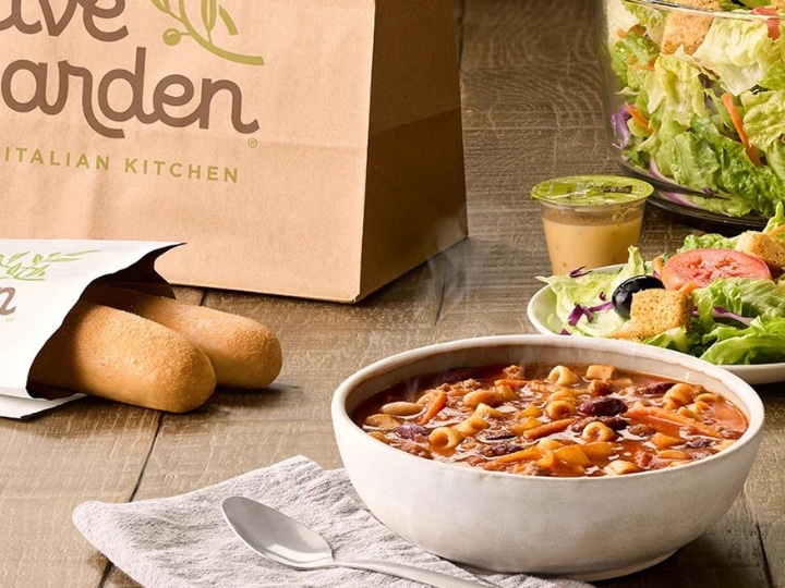 A bowl of soup, part of a plate of salad, and part of a bag of breadsticks on a wood table.