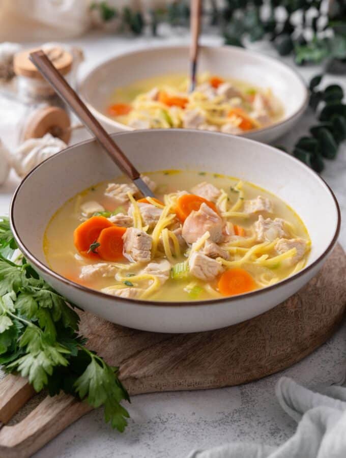 A bowl of turkey noodle soup with chunks of carrots and celery. There is a spoon in the bowl and a second bowl in the background.