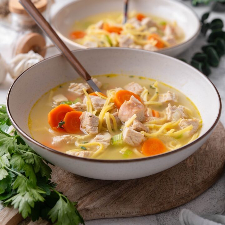 A bowl of turkey noodle soup with chunks of carrots and celery. There is a spoon in the bowl and a second bowl in the background.