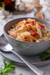 A bowl of cabbage soup with chunks of tomato, carrots, and seasonings. The bowl is on a plate next to a spoon.