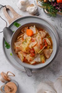 Overhead view of a bowl of cabbage soup with chunks of carrots, tomatoes, and seasonings. The bowl is on top of a plate next to a spoon and sprigs of cilantro.