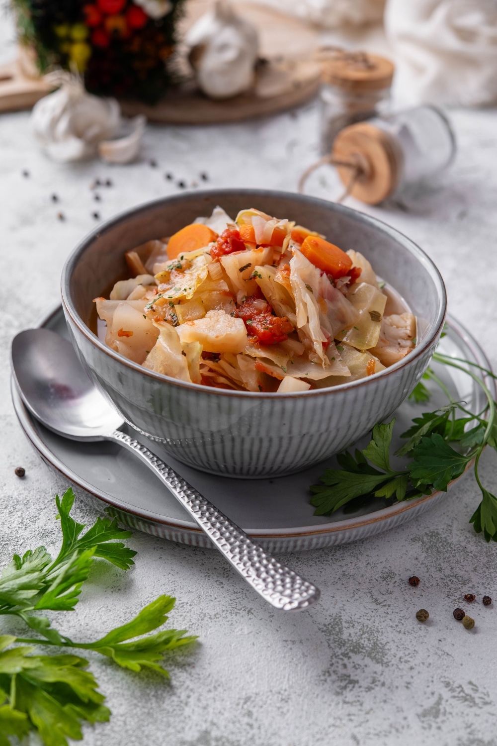 A bowl of cabbage soup with chunks of tomato, carrots, and seasonings. The bowl is on a plate next to a spoon and sprigs of cilantro.