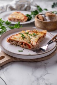 Two slices of crustless pizza garnished with fresh parsley on a white ceramic plate. The plate is on a wooden serving board and has two forks. There is a second plate of crustless pizza and a wooden bowl in the background.