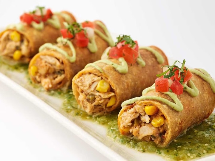 Four cheesecake factory Chicken Taquitos on top of green salsa on a white plate.