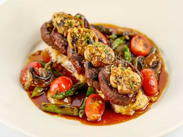 Five rounds f cheesecake factory Grilled Steak Medallions on a top of a white plate with cut tomatoes and asparagus.