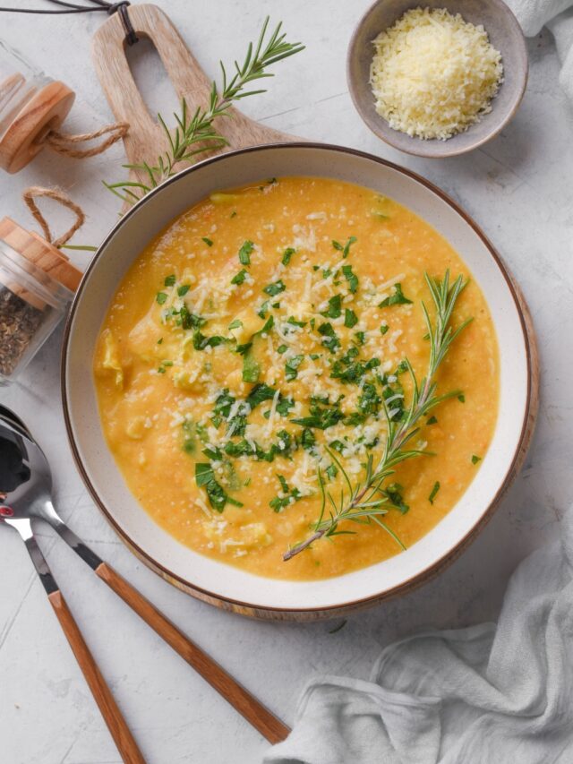 Overhead view of a bowl of creamy soup with parmesan cheese, rosemary, and kale on top. Two spoons and a bowl of parmesan cheese are next to the soup.