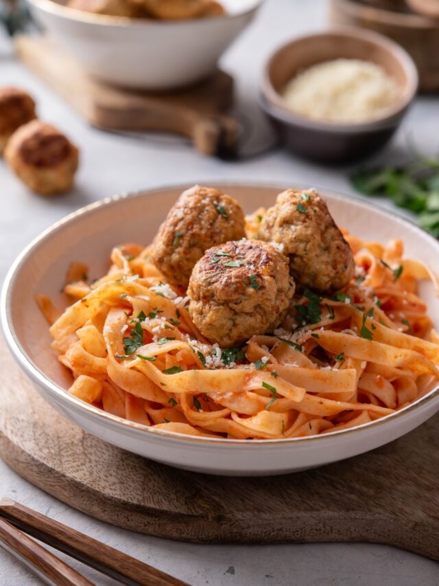 A plate of pasta in red sauce with three meatballs on top. The plate is on a wooden board next to a set of silverware.