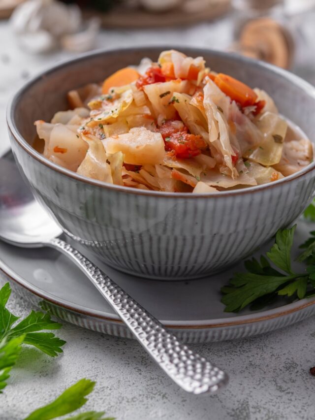A bowl of cabbage soup with chunks of tomato, carrots, and seasonings. The bowl is on a plate next to a spoon.